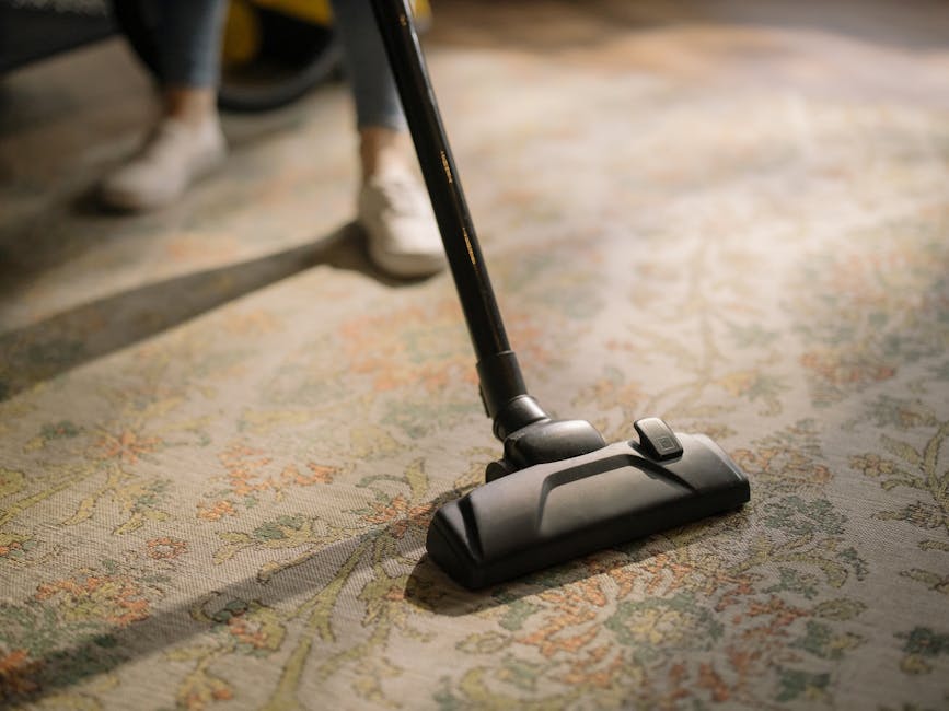 A person vacuuming a dark grey carpeted floor in a large, dimly lit room, possibly a conference or screening room, with a white projection screen on the wall behind them. The individual, dressed in ca