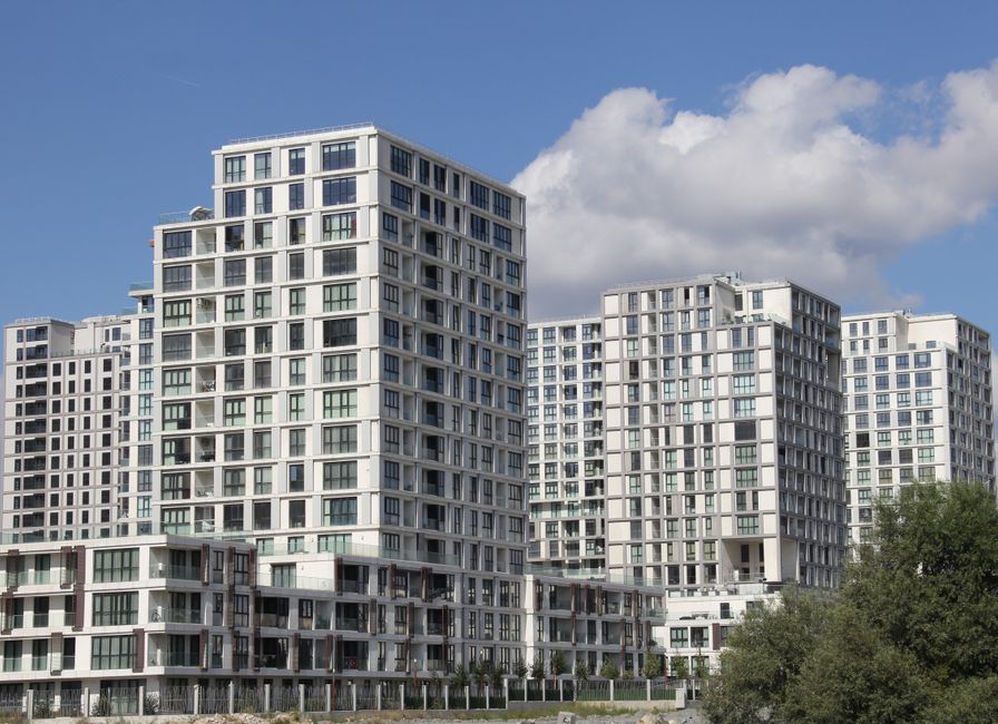 A modern residential complex consisting of multiple high-rise buildings with white facades and large glass windows, set against a blue sky with scattered clouds. The buildings feature a geometric desi